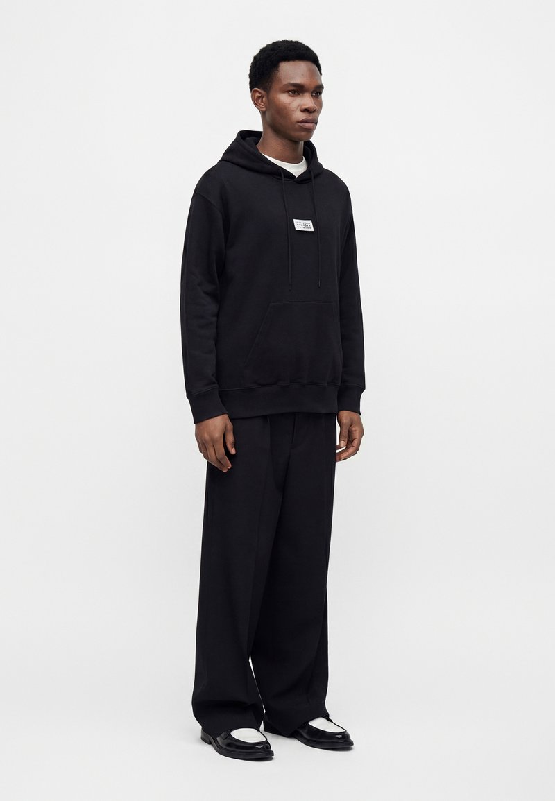 Jeune homme debout portant un sweat à capuche noir avec poche avant, un pantalon noir à jambes larges et des chaussures noires et blanches sur un fond blanc uni.