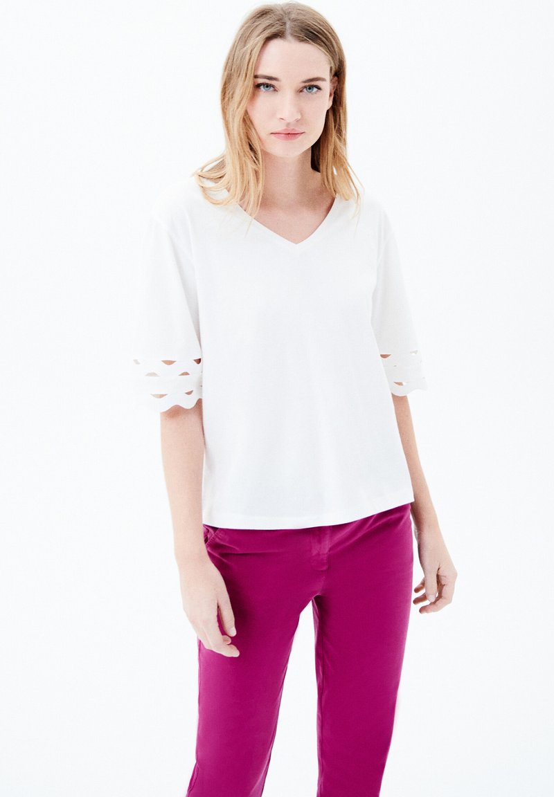 Top bianco a scollo a V con dettagli a scallop sulle maniche, abbinato a pantaloni rosa brillante. Tessuto liscio, vestibilità comoda, senza motivi visibili.
