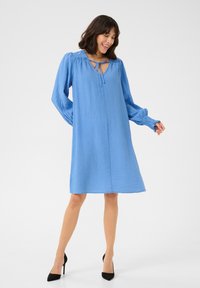 Robe bleu clair en tissu texturé, dotée d'un col à nouer et de longues manches ballon. Détails en fronces aux épaules et à l'ourlet.