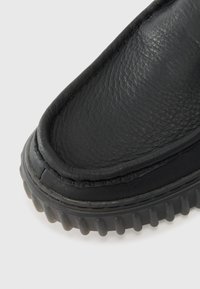 Black slip-on shoe featuring a smooth leather upper, rounded toe, and raised rubber sole with textured grip. Stitched detailing along the edge.