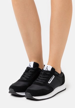 Baskets Calvin Klein noir et blanc portées aux pieds, avec lacets et écussons logo sur les languettes et les côtés, posées sur une surface blanche.