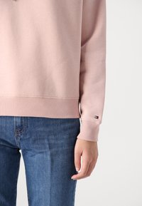 Sweatshirt rose clair avec ourlet et poignets côtelés, arborant une petite étiquette logo sur la manche. Assorti à des jeans en denim bleu.