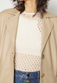 Veste en cuir beige avec un col cranté portée sur un haut en tricot texturé blanc à motif à pois et une taille de couleur claire. Un jean en denim est visible.