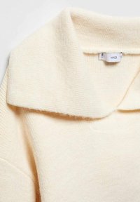 Pull couleur crème avec une texture côtelée et un col rabattable. L'étiquette indique "MNG". Le tissu semble doux et chaud.