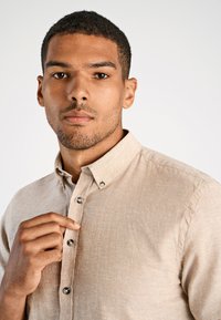 Lichtbeige knoopshirt gemaakt van textuurstof. Heeft een kraag en contrasterende knopen, met een close-up van de halslijn.