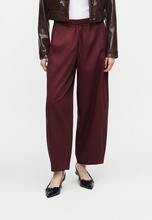 Γυναίκα που φοράει μπορντό παντελόνι με φαρδιά μπατζάκια, μαύρα μυτερά slingback παπούτσια με λεπτομέρεια φιόγκο, λευκή μπλούζα και κοντό καφέ δερμάτινο μπουφάν.