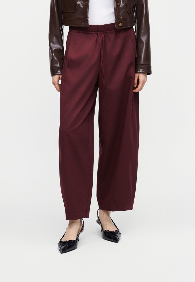 Γυναίκα που φοράει μπορντό παντελόνι με φαρδιά μπατζάκια, μαύρα μυτερά slingback παπούτσια με λεπτομέρεια φιόγκο, λευκή μπλούζα και κοντό καφέ δερμάτινο μπουφάν.