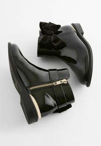 Schwarze Lack-Ankle-Boots mit glänzender Oberfläche, versehen mit einer hinteren Samtschleife, Reißverschluss und einem niedrigen, gestapelten Absatz.