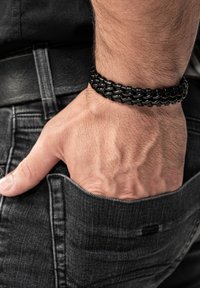 Black metal chain bracelet featuring a braided design, resting on a hand with a textured wrist against dark denim. Pocket and belt visible.