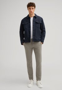 Giacca di jeans blu navy con due tasche sul petto, abbinata a pantaloni grigio chiaro e sneakers bianche. La texture è liscia e il design è informale.
