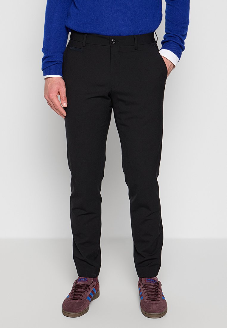 Schwarze Hosen mit schlanker Passform, Fronttaschen und Knopfverschluss. Getragen mit einem blauen Pullover und burgunderfarbigen Sneakers mit blauen Streifen.
