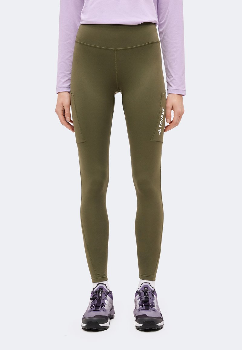 Personne portant des leggings moulants vert olive avec poches latérales et des chaussures de randonnée violettes sur un fond clair uni.