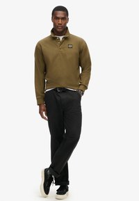 Olivgrünes Langarm-Polo-Shirt mit Kragen, besticktem Logo, kombiniert mit schwarzen Hosen und schwarzen Sneakers mit weißen Sohlen.