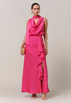 Frau trägt ein ärmelloses, leuchtend pinkfarbenes Satin-Kleid mit Rüschen und hohem Beinschlitz, goldenen Armreif, Ohrringe, Clutch und hautfarbene High Heels.