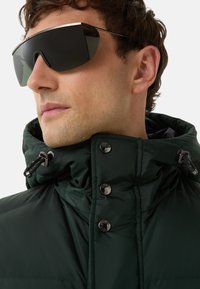 Grøn pufjakke med høj krave, prydet med sølvfarvede trykknapper og snor. Model iført mørke, oversized solbriller.