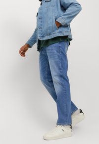 Vaaleansininen farkkutakki, jossa taskut, kerrospukeutunut tummanvihreän paidan kanssa, yhdistetty vaaleansinisiin farkkuihin ja valkoisiin tennareihin, joissa on mustia yksityiskohtia.