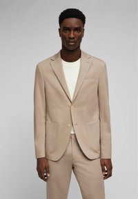 Mann in beigem Anzug mit zweiknöpfigem Blazer und passender Hose, kombiniert mit einem weißen Rundhals-T-Shirt, vor einem schlichten grauen Hintergrund stehend.