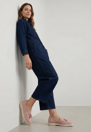 Kvinde, der læner sig op ad en væg iført en mørk denim-dragtdragt og lyserøde loafers med kvaster, stående på en lysegrå gulv.