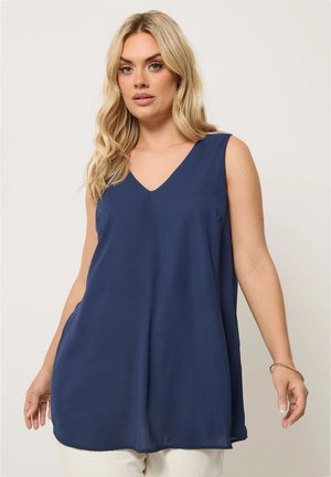 Femme blonde portant un haut sans manches bleu marine à col en V et un pantalon blanc, debout devant un fond clair uni.