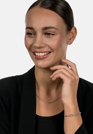 Schmuck in Silberoptik mit einem verlinkten Kettendesign. Das Model trägt eine Halskette und ein Armband mit geometrischen Formen und glatter Textur.