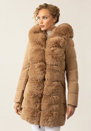 Frau in einem beigen Steppmantel mit einem großen, flauschigen Pelzkragen, die vor einem schlichten hellen Hintergrund steht und zur Seite schaut.