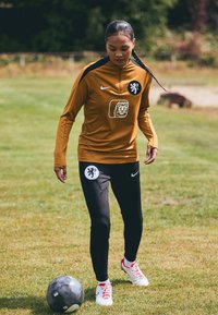 Nike tracksuit in orange and black with lion emblem; black pants; player dribbling a soccer ball on grass. White shoes with red accents.