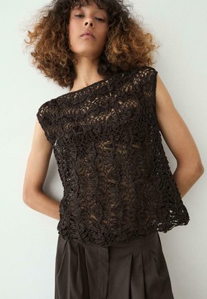 Frau mit lockigem Haar, die ein ärmelloses, schwarzes, locker gestricktes Top über nackter Haut und dunkle, plissierte Hosen vor einem einfarbigen Hintergrund trägt.