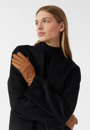 Braune Lederhandschuhe mit glatter Textur, ausgestattet mit vier länglichen Fingerabschnitten, kombiniert mit einem dunklen schwarzen Wollmantel.