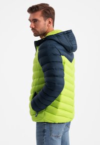 Vuorattu takki hupulla, jossa on vihreä vartalo ja tummansiniset hihat. Teksturoitu, tikattu muotoilu sivutaskuilla ja vetoketjukiinnityksellä.