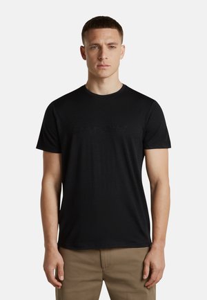 Mann mit kurzen braunen Haaren trägt ein schlichtes schwarzes T-Shirt mit kurzen Ärmeln und beige Hose, steht mit neutralem Gesichtsausdruck vor weißem Hintergrund.