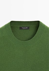 Zielony sweter z okrągłym dekoltem i prążkowanym kołnierzem, z czarną metką wszytą wewnątrz z białym napisem.