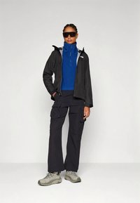 Femeie îmbrăcată în echipament de drumeție, cu geacă neagră, bluză albastră cu fermoar, pantaloni cargo negri, încălțăminte de drumeție bej și ochelari de soare mari, negri.