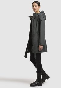 Donkergrijze waterdichte parka met een capuchon, rits- en knoopsluiting, zijzakken en een gestructureerde binnenvoering, gedragen over zwarte leggings en laarzen.