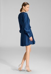 Vrouw draagt een marineblauwe jurk met lange mouwen en zilveren sandalen met hoge hak, staand met haar lichaam iets zijwaarts gedraAid.