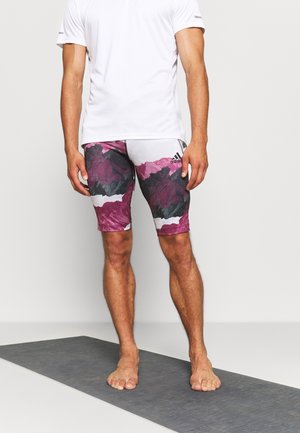 Mężczyzna stojący boso na szarej macie, ubrany w białą koszulę i czarne, fioletowe oraz białe sportowe szorty marki Adidas z wzorem.