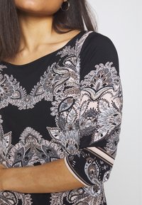 Černé šaty s květinovým vzorem v odstínech šedé a růžové; mají výstřih do „srdce“ a tříčtvrteční rukávy s akcentními pruhy.