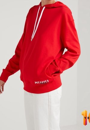 Personne portant un sweat à capuche rouge vif avec des cordons blancs et un pantalon blanc, debout avec une main dans la poche du sweat à capuche sur un fond uni.