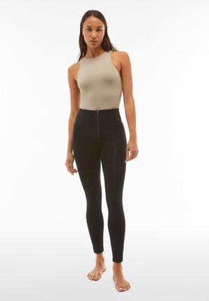 Leggings neri a vita alta con cerniera frontale, abbinati a un top aderente beige. Tessuto liscio senza motivi visibili.