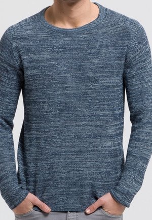 Homme portant un pull en maille bleu chiné ajusté à manches longues, mains dans les poches, debout devant un fond uni.