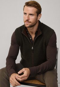 Tmavě zelená pletená vesta se zipem vpředu, nošená přes hnědou košili s dlouhým rukávem. Vesta má žebrovanou strukturu a límeček.