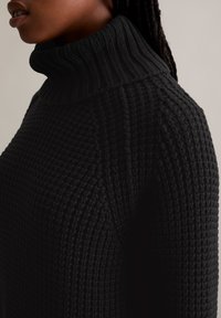 Schwarzer Strickpullover mit hohem Rippenkragen, strukturiertem Muster und taillierter Passform. Nahaufnahme, die sich auf die Details von Kragen und Schulter konzentriert.