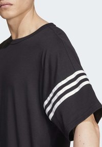 Man som bär en svart kortärmad skjorta med tre vita ränder på överarmen, avskuren vid halsen och axeln.