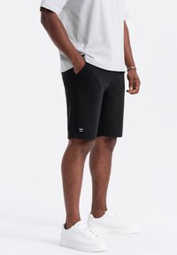 Shorts noirs texturés avec un design côtelé, poches latérales et une petite étiquette de marque. Assortis à des baskets blanches et une chemise à manches courtes grise.