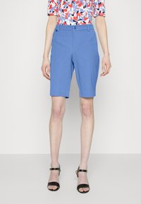 Kvinna i blå knälånga shorts, flerdelad blommig topp och svarta klacksandaler med öppen tå som står mot en enfärgad bakgrund.