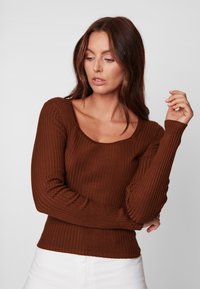 Pull marron à manches longues côtelé avec un col arrondi et une coupe ajustée. Texture lisse et forme structurée, associé à un pantalon blanc taille haute.