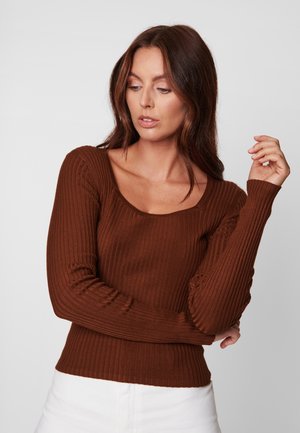 Pull marron à manches longues côtelé avec un col arrondi et une coupe ajustée. Texture lisse et forme structurée, associé à un pantalon blanc taille haute.