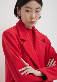 Femme avec les cheveux courts et foncés, portant un manteau rouge et un pull, des boucles d'oreilles en argent en forme de hoop et plusieurs bagues en argent, les bras croisés, expression neutre.