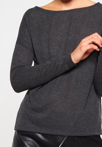 Maglia lunga grigia scura con scollo ampio e maniche aderenti. Il tessuto appare morbido e leggero, leggermente testurizzato.