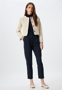 Chaqueta corta crema con botones dorados, combinada con pantalones de cintura alta en color azul marino. Modelo de pie, con las manos en los bolsillos. Textura de tela suave.