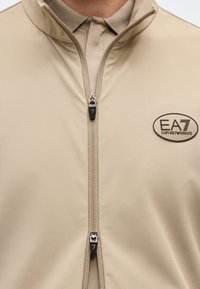 Μπεζ μπουφάν με φερμουάρ και δύο μαύρα και ασημένια φερμουάρ, με το λογότυπο EA7 Emporio Armani στο αριστερό στήθος, φορεμένο πάνω από μπέζ πόλο μπλουζάκι.